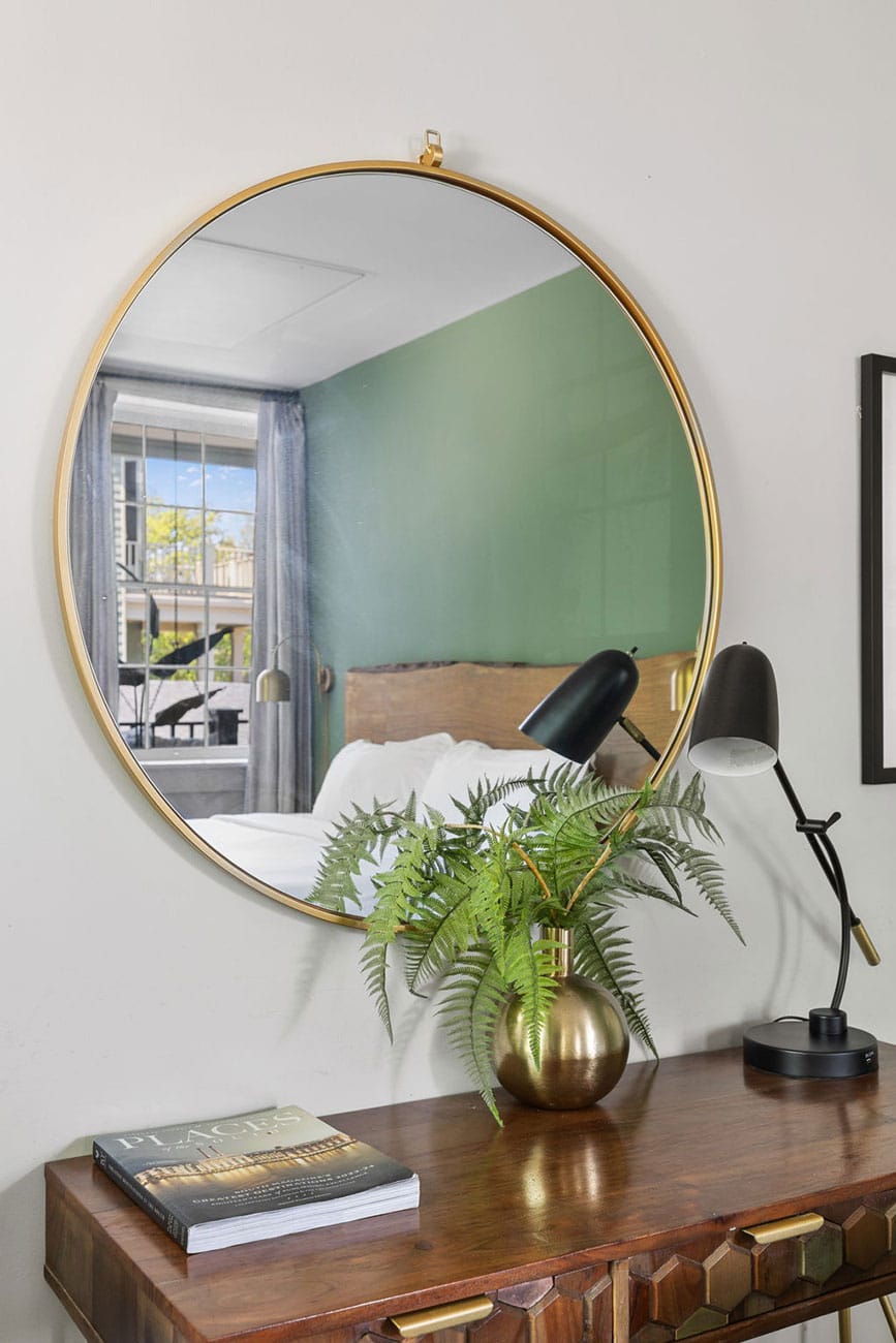 Round gold-framed mirror above a wooden console table with a brass fern vase and a black desk lamp; a magazine rests on the table.