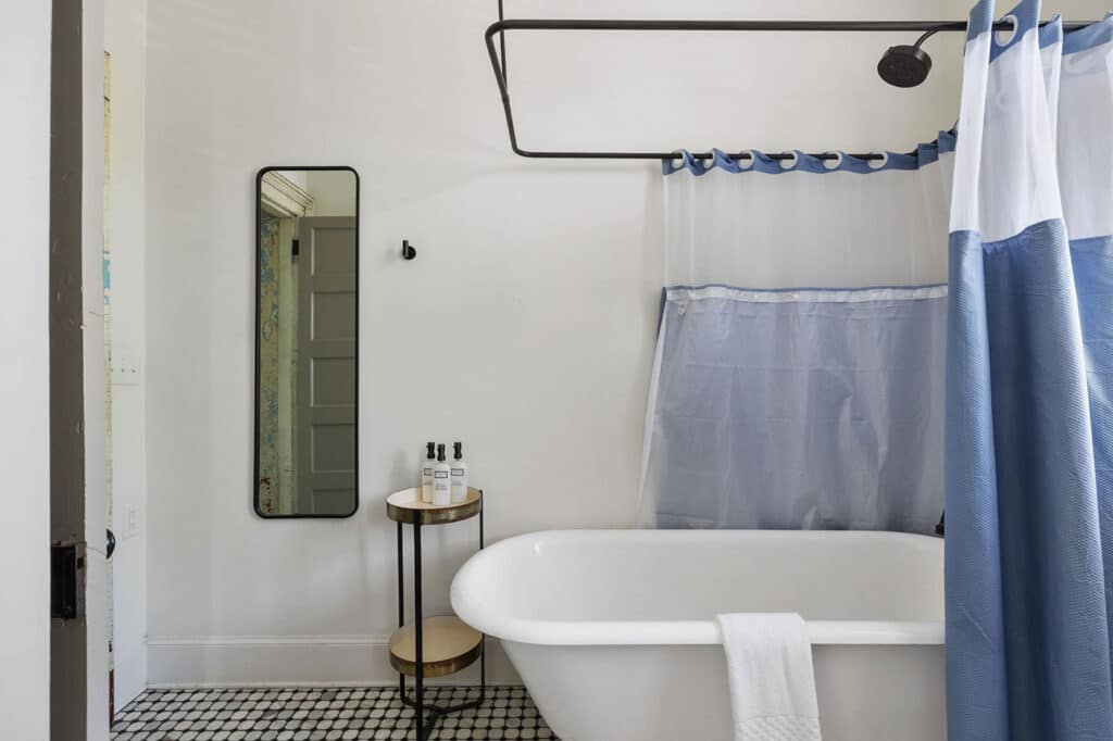 White bathroom with a freestanding tub, blue-and-white shower curtain, and a small round side table with bath products beside it.