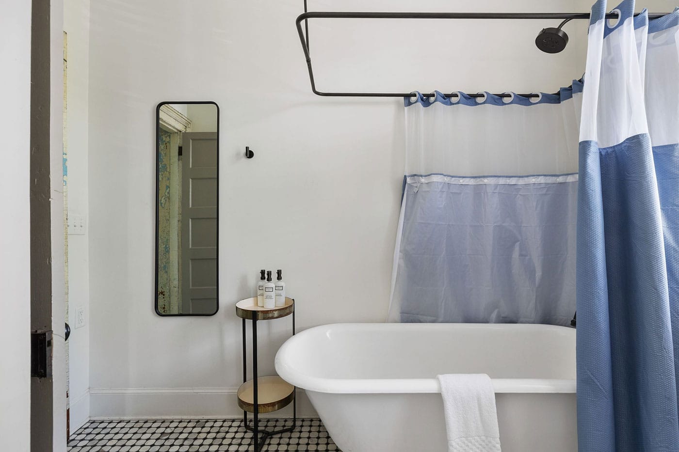 White bathroom with a freestanding tub, blue-and-white shower curtain, and a small round side table with bath products beside it.
