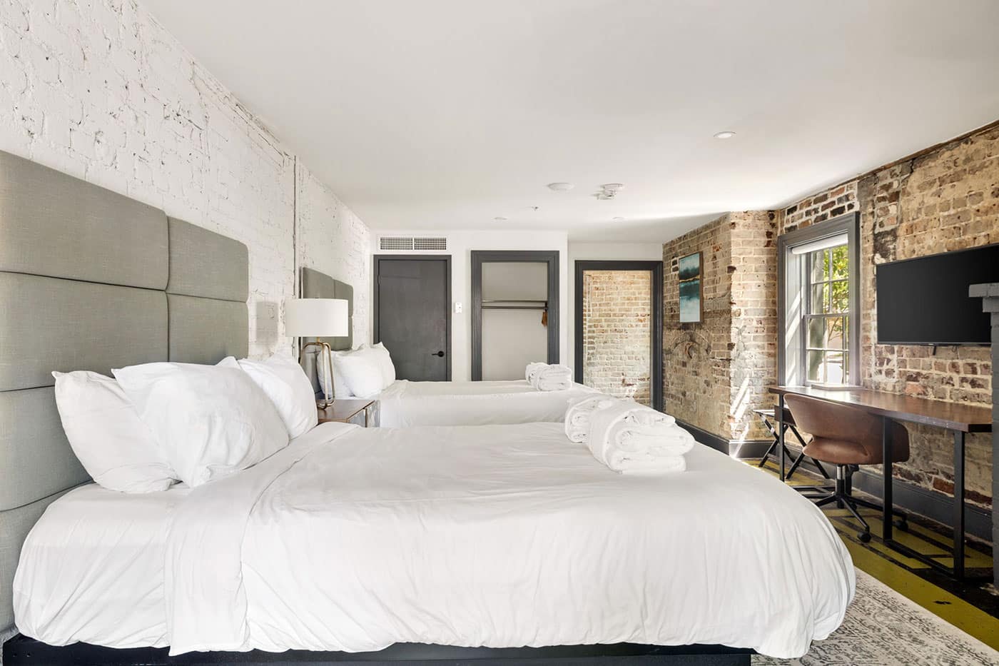 Industrial-style hotel room with two large white beds, gray padded headboards, and folded towels on the beds beside a wooden desk and chair near a brick wall window.