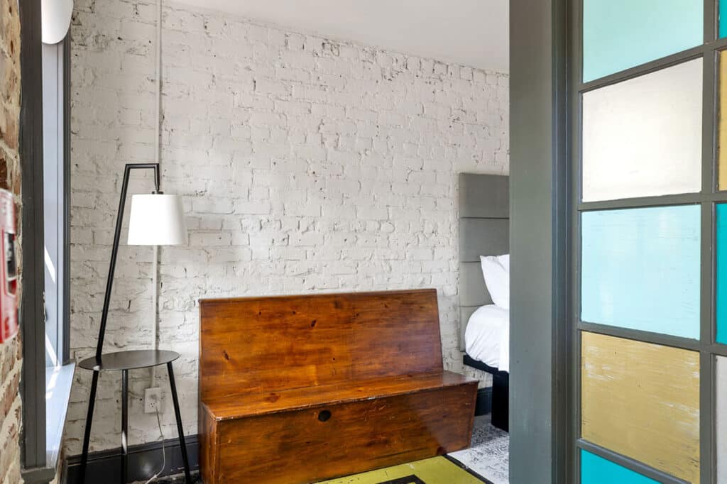 A rustic bedroom corner with a wooden storage bench, a black floor lamp, and a white brick wall beside a bed partially visible at the back; colorful stained-glass door on the right.