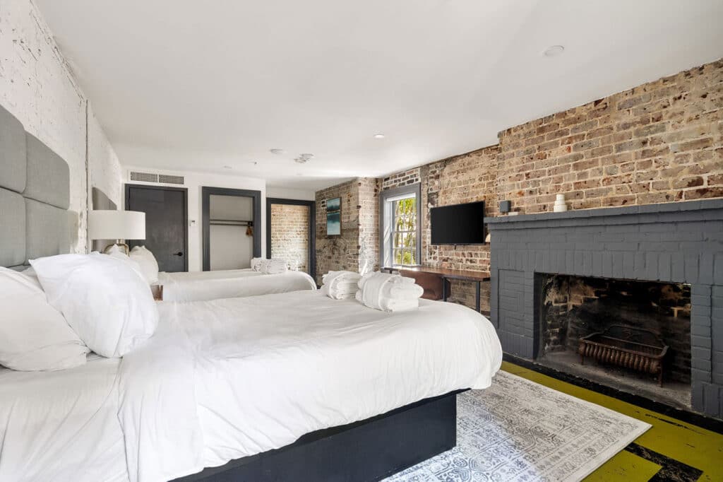 Two double beds with white linens in a brick-walled loft hotel room, featuring a gray fireplace and a wall-mounted TV.