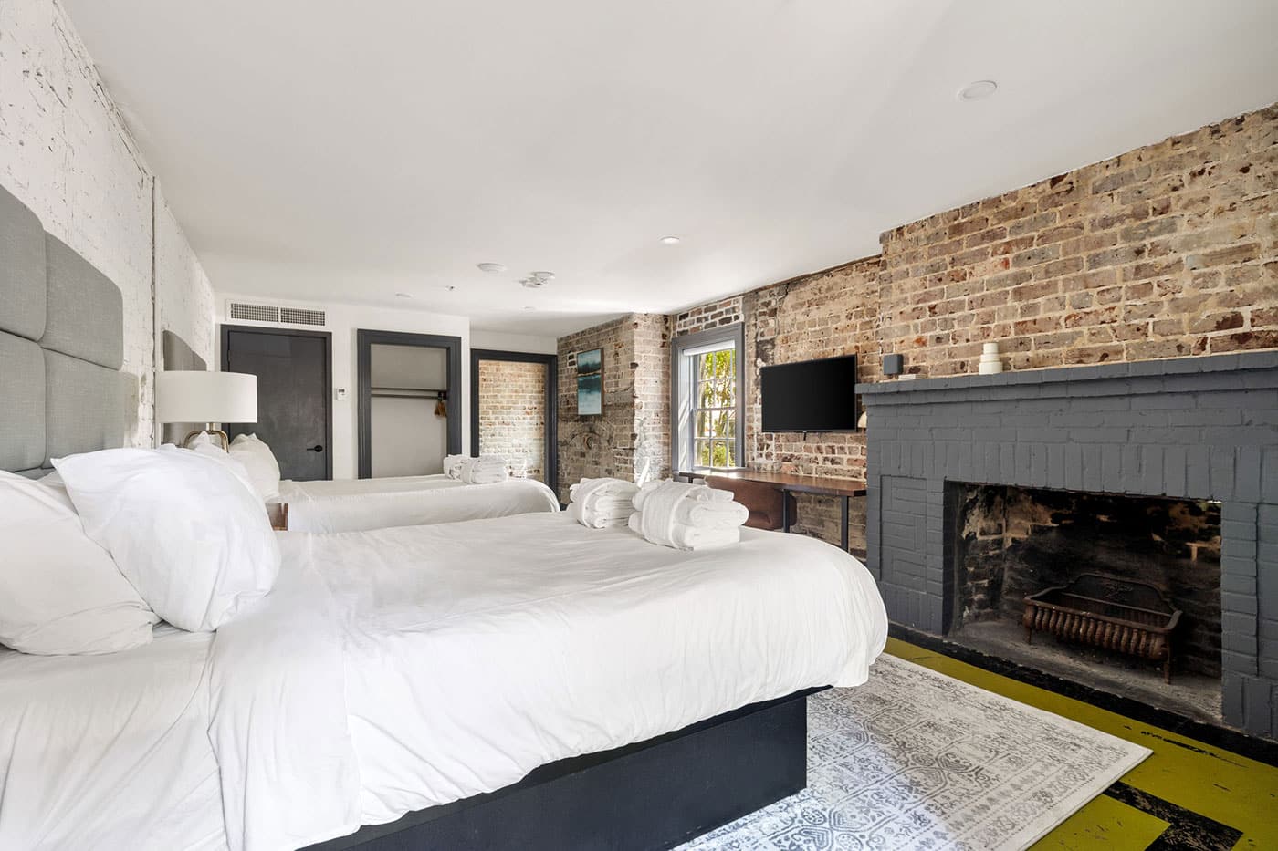Two double beds with white linens in a brick-walled loft hotel room, featuring a gray fireplace and a wall-mounted TV.