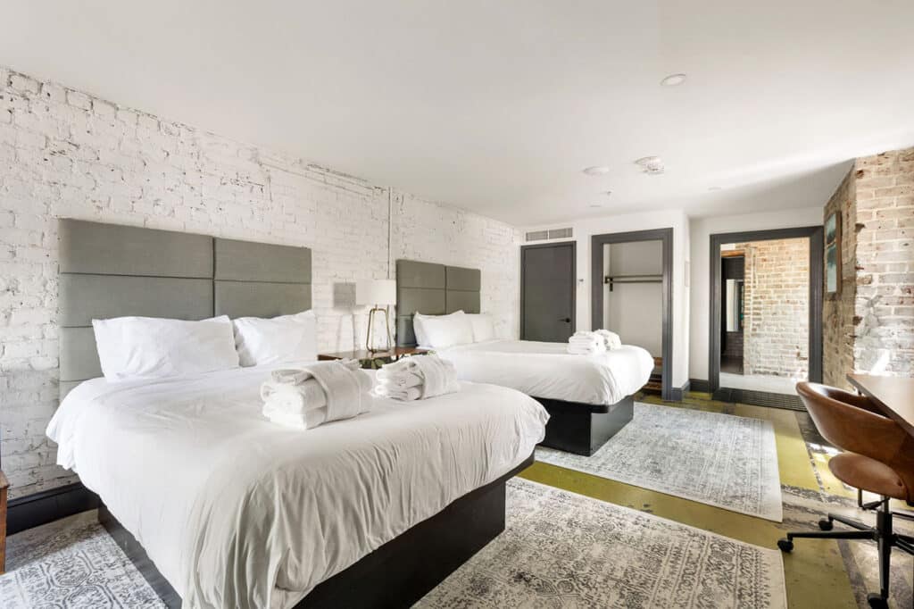 Hotel room with two queen beds, white linens, and gray upholstered headboards against white brick walls, towels folded on the beds.