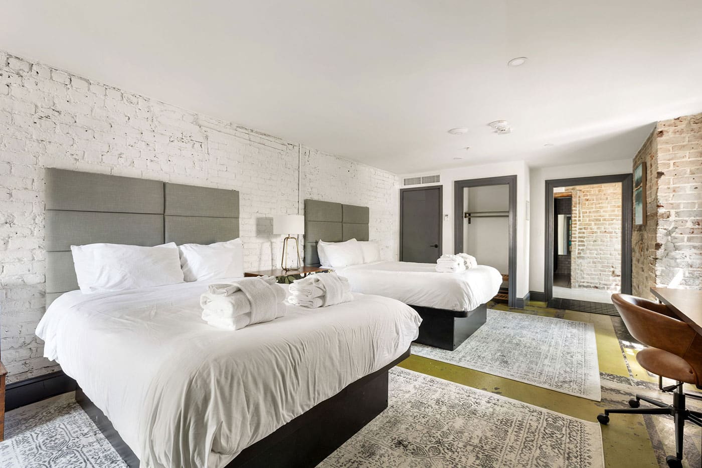 Hotel room with two queen beds, white linens, and gray upholstered headboards against white brick walls, towels folded on the beds.