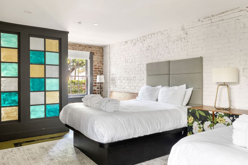 Bright bedroom with white brick wall, two white beds, gray upholstered headboard, and a nightstand lamp; colorful glass-pane door on the left.