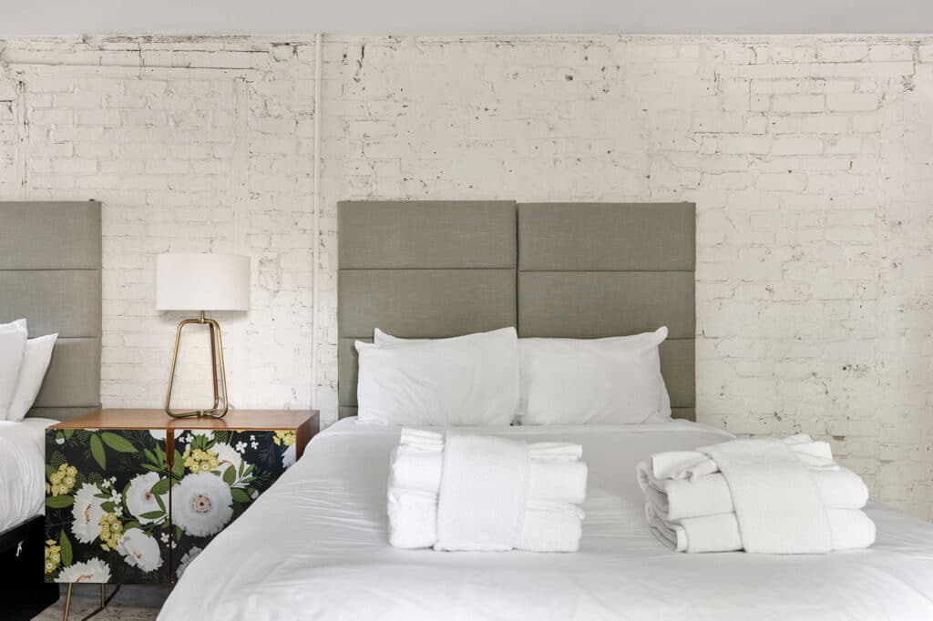 Contemporary hotel bedroom with gray upholstered headboard, white bedding, and folded towels on the bed; floral nightstand and white brick wall backdrop.