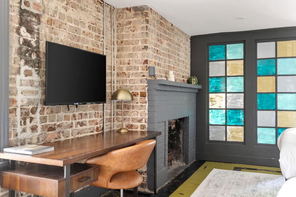 Home office with exposed brick wall, mounted TV, wooden desk, brass lamp, and a gray brick fireplace beside a stained glass window paneling.