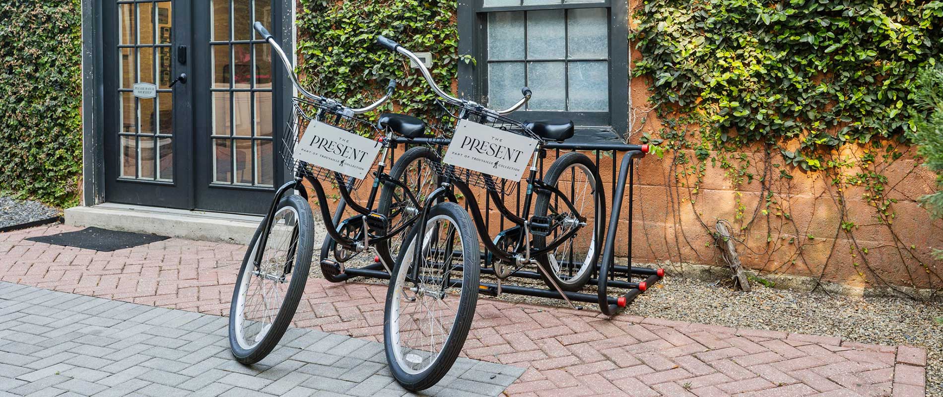 Two black bicycles locked to a rack in front of a ivy-covered brick building; signs on the bicycles read 'THE PRESENT'.